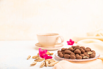 Almond in chocolate dragees on ceramic plate and a cup of coffee on white concrete background. Side view, selective focus, copy space.