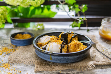 Fried white ice cream on blue plate, grey table with flowers near window. Natural day light in the kitchen. Homemade dessert, rustic or country style. Copy space. Hot ice in corn breading.