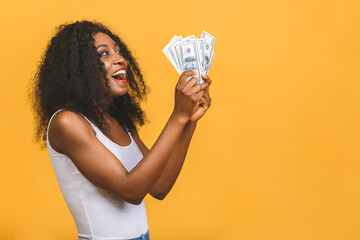 Happy winner. Portrait of african american successful woman 20s with afro hairstyle holding lots of money dollar banknotes isolated over yellow background.