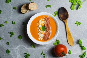 Salmorejo dish with eggs and ham, accompanied by tomato and bread, along with a black cloth and a wooden spoon on a gray stone.