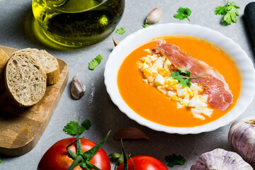 Salmorejo dish with eggs and ham, accompanied by tomato, oil, garlic and bread, next to a black cloth and on a gray stone.