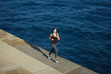 Full length portrait of a young woman with fitness body running at the coastline next to the ocean, athletic girl jogging over amazing big waves background at sunny day