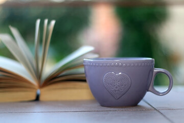 Purple cup of tea or coffee and open book in a garden. Selective focus.