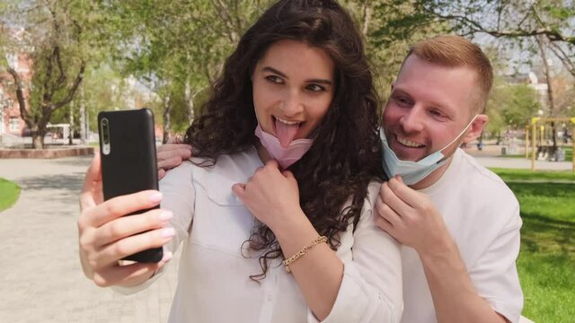 Slowmo dolly-in shot of happy young woman and man taking of face masks and posing together for selfie outdoors on sunny summer day