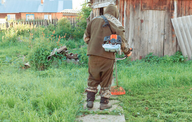 Asian man in a mosquito suit with a professional brush cutter trimmer mows the grass in the yard. green lawn, old fence of the village house. Sunny summer weather. cutting  lawn mower.