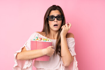 Teenager girl over isolated pink background with 3d glasses and holding a big bucket of popcorns