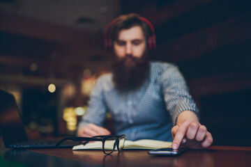 Fototapeta na wymiar Select focus on desktop with stylish optical eyeglasses with black frame and man's finger touch screen of modern telephone.Bearded man in headphones sitting with laptop on blurred background