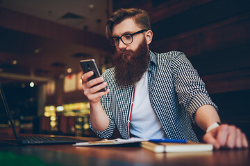 Smart bearded student in optical eyeglasses reading new post on internet website during break using modern telephone device sitting at desktop with digital netbook and books in stylish coworking space
