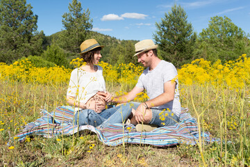 Pregnant and smiling couple sitting on a blanket in a meadow full of tall yellow flowers. Husband touching the pregnant belly of his wife.  