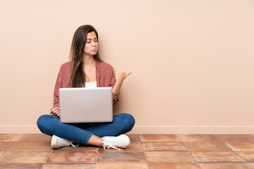 Teenager student girl sitting on the floor with a laptop unhappy and pointing to the side