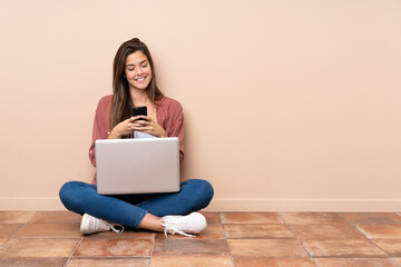 Teenager student girl sitting on the floor with a laptop sending a message with the mobile