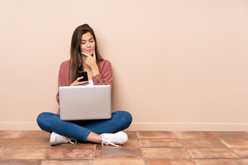 Teenager student girl sitting on the floor with a laptop thinking and sending a message