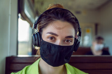 A young girl in a protective mask and headphones looks at the camera. A brunette in a protective mask listens to music and rides the train