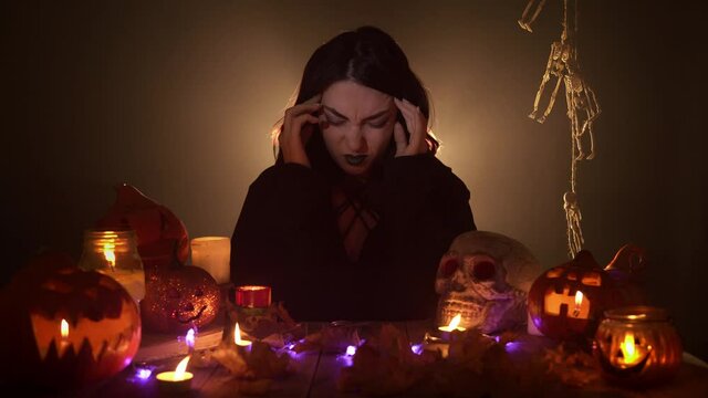 Young woman in black mantle is in pain with terrible migraine headache. The woman is sitting among different halloween decorations. Theme of revealing feelings and emotions.