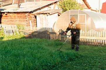 Asian man in a mosquito suit with a professional brush cutter trimmer mows the grass in the yard....