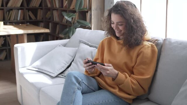 Happy young hispanic latin ethnic teen girl sit on sofa at home holding phone looking at screen watching social media video content, movie or stream online in mobile app laughing using smart phone.