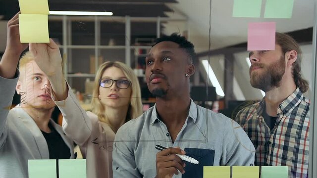 Close up of handsome contented three multiracial men and one pretty light-haired female colleague which working with new business strategy and modeling it on glass wall
