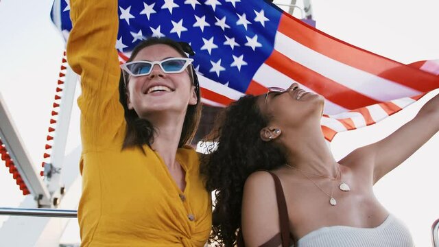 Young mixed race girls are laughing and dancing, holding flag of USA above their heads and waving it while sitting in a ferris wheel booth. Slow motion