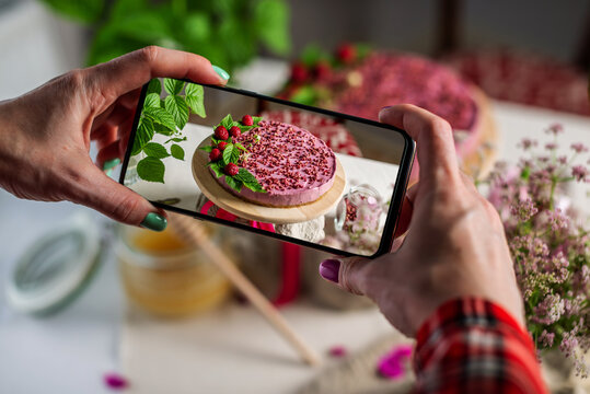 Woman is taking photos on a mobile phone camera of a beautiful and appetizing mousse cake decorated with berries and green leaves