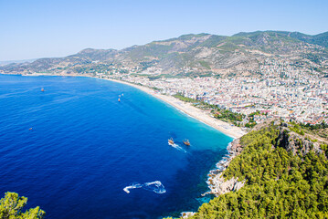 City on the coast between the mountains and blue sea with a sandy beach. View on the Kleopatra Beach, Alanya, Turkey. Mediterranean Sea. Tourist coastal town with cute houses near the sea with boats.