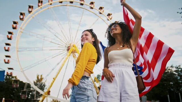 Young mixed race girls in sunglasses are smiling and dancing, holding flag of USA and waving it, posing in park against a ferris wheel. Summer day. 4th of July