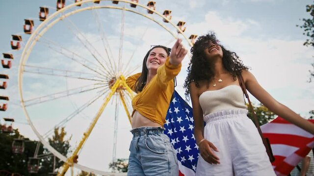 Young mixed race ladies in sunglasses are smiling, holding flag of USA above their heads and waving it while dancing in park against a ferris wheel. Slow motion - Powered by Adobe