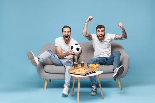 Excited men guys friends in white t-shirt sit on couch isolated on blue background. Sport leisure concept. Cheer up support favorite team with soccer ball scream expressive gesticulating with hands.