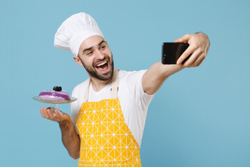 Excited young bearded male chef cook baker man in apron white t-shirt toque chefs hat isolated on blue background studio. Cooking food concept. Hold dessert cake doing selfie shot on mobile phone.
