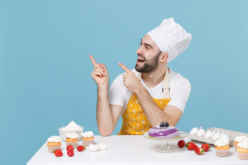Cheerful bearded male chef or cook baker man in apron white t-shirt toque chefs hat cooking at table isolated on blue background. Cooking food concept. Mock up copy space Point index fingers aside up.