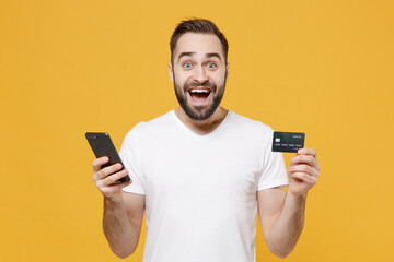 Excited young bearded man guy in white casual t-shirt posing isolated on yellow wall background studio portrait. People lifestyle concept. Mock up copy space. Hold credit bank card using mobile phone.
