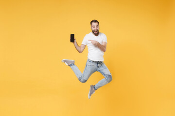 Excited young bearded man guy in white casual t-shirt isolated on yellow background. People lifestyle concept. Mock up copy space. Jumping point index finger on mobile phone with blank empty screen.