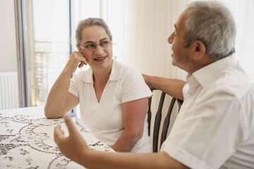 Old aged, Turkish, happy senior couple enjoying time together, looking to each other, laughing, sitting in living room on chair.