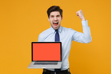 Screaming young business man in classic blue shirt tie isolated on yellow background. Achievement career wealth business concept. Hold laptop pc computer with blank empty screen doing winner gesture.