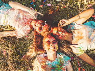 Three happy beautiful girls making party at Holi colors festival in summer time.Young  smiling women friends having fun after music event at sunset.Models lying on the grass in sunglasses.