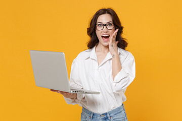 Excited young business woman in white shirt glasses isolated on yellow background studio. Achievement career wealth business concept. Mock up copy space. Hold laptop pc computer, put hand on cheek.