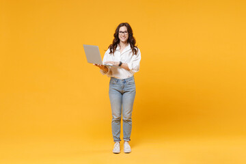 Smiling young brunette business woman in white shirt glasses isolated on yellow background in studio. Achievement career wealth business concept. Mock up copy space. Working on laptop pc computer.