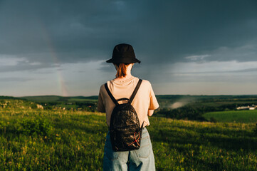 Woman hiker in stylish casual clothes and backpack stands with her back to a beautiful mountain...