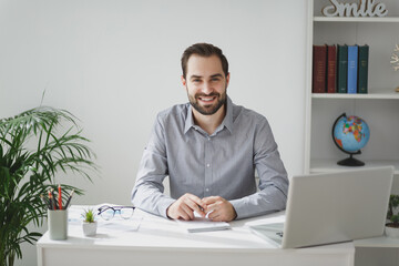 Smiling attractive young bearded business man in gray shirt sitting at desk working on laptop pc computer in light office on white wall background. Achievement business career concept. Looking camera.