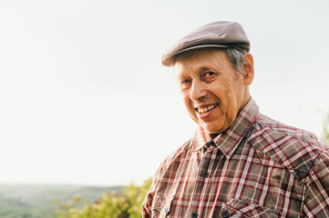 Naklejka na ściany i meble Portrait of old happy man in cap and shirt outdoors at sunset, looking at camera and smiling. Retired man posing at camera, close up photo. Portrait of an old positive grandfather.