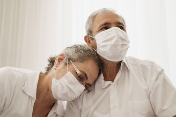 Sad looking old couple wearing masks. Wife puts her head on her husband's shoulder