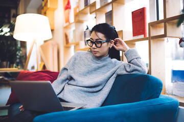 Female Asian student sitting in cozy lounge working on laptop