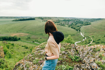 The back of a stylish hipster girl stands on a hill and looks at the beautiful landscape from the top of the mountain, standing on a rock. Woman tourist enjoys the landscape from the mountain Tovtry
