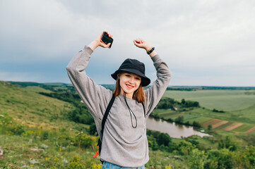 Happy hipster girl in casual clothes stands on a hill with a smartphone in hand and rejoices with raised hands on the background of a beautiful Ukrainian landscape with a lake.Travel to Ukraine.Tovtry