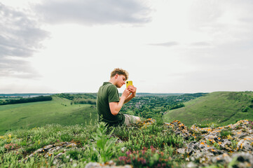 Naklejka na ściany i meble Young man in casual clothes rocks on the mountain on the background of a beautiful Ukrainian landscape, takes photos on a smartphone camera. tourist rests sitting on a rock of a hill, uses smartphone