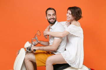 Side view of smiling young couple two friends guy girl in white summer clothes driving moped isolated on orange wall background studio. Driving motorbike transportation concept. Mock up copy space.