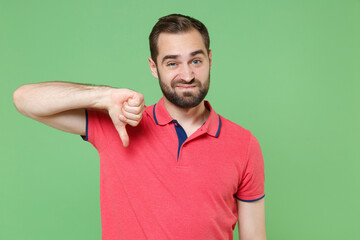 Displeased young bearded man guy in casual red pink t-shirt posing isolated on green wall background studio portrait. People sincere emotions lifestyle concept. Mock up copy space. Showing thumb down.