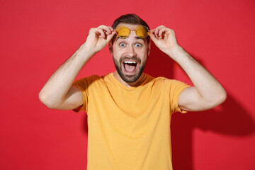 Excited young bearded man guy in casual yellow t-shirt glasses posing isolated on red background studio portrait. People sincere emotions lifestyle concept. Mock up copy space. Keeping mouth open.