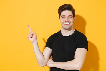 Fototapeta premium Smiling young man guy 20s in casual black t-shirt posing isolated on yellow background studio portrait. People sincere emotions lifestyle concept. Mock up copy space. Pointing index finger aside up.