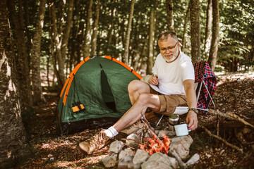 Mature man sitting in front of a tent and near campfire and writing in a notebook.