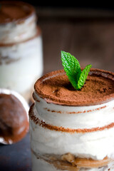 Close up of a jar filled with tiramisu with cocoa powder on top and mint leaves as decoration and a second vase on the background. Dark food concept.